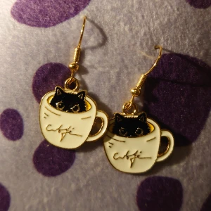Örhängen med kattmotiv i kaffekopp - Charmiga örhängen med en svart katt som tittar upp ur en vit kaffekopp. Perfekta för kattälskare och de som gillar unika accessoarer. Hängena har en guldig krok och är både lekfulla och stilrena.