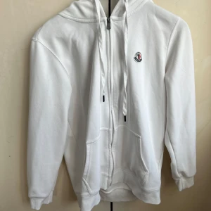 Vit hoodie från Moncler - Säljer en stilren vit hoodie från Moncler med dragkedja och justerbar huva. Tröjan har ett litet broderat Moncler-emblem på bröstet och ribbade muddar vid ärmslut och nederkant. Perfekt för en avslappnad look.