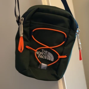 Grön axelväska från The North Face - Snygg grön axelväska från The North Face med orange detaljer och dragkedja. Perfekt för att bära med sig det nödvändigaste. Justerbar axelrem för bekväm passform.