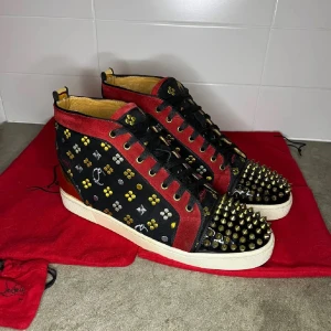 Svarta sneakers med nitar från Christian Louboutin - Unika svarta sneakers med röda detaljer och guldfärgade nitar från Christian Louboutin. Skorna har en mönstrad yta med snörning och en ikonisk röd sula. Perfekta för den som vill sticka ut med stil.