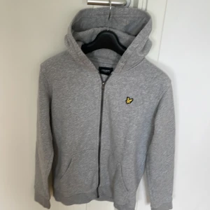 Grå hoodie från Lyle & Scott - Säljer en stilren grå hoodie från Lyle & Scott med dragkedja och deras ikoniska logga på bröstet. Perfekt för en avslappnad look. Den har långa ärmar och en praktisk huva.