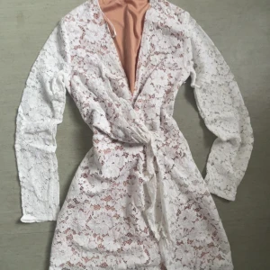 Vit spetsklänning med knytband - Elegant vit spetsklänning med långärmad design och knytband i midjan. Klänningen har en omlottstil och är perfekt för speciella tillfällen som studenten. Den har en ljus persikofärgad underklänning som ger en fin kontrast till den vita spetsen.