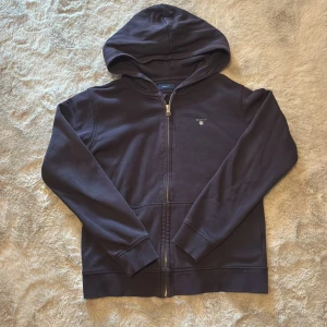 Mörkblå hoodie med dragkedja från Gant - Säljer en stilren mörkblå hoodie från Gant med dragkedja och huva. Tröjan har långa ärmar och en liten logotyp på bröstet. Perfekt för en avslappnad stil. Stl 158