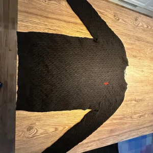 Svart stickad tröja från Ralph Lauren - Säljer en stilren svart stickad tröja från Ralph Lauren med kabelstickat mönster. Tröjan har långa ärmar och en liten röd logotyp på bröstet. Perfekt för kyligare dagar! 