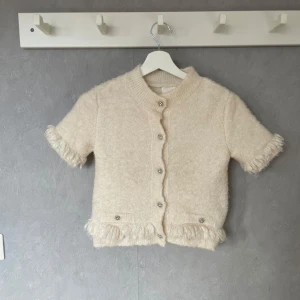 Beige kortärmad kofta med fransar - Charmig beige kofta med korta ärmar och dekorativa fransar vid ärmslut och nederkant. Koftan har knappar framtill och en mjuk, mysig textur. Perfekt för en stilren och bekväm look.