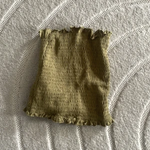 Olivgrön smockad tubtopp - Säljer en olivgrön tubtopp med smockad design och volangdetaljer upptill och nedtill. Perfekt för sommardagar och kan enkelt matchas med olika outfits. Passar bra för en avslappnad och stilren look.