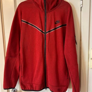 Röd hoodie från Nike - Snygg röd hoodie från Nike med dragkedja och huva. Den har svarta och vita ränder över bröstet och en praktisk ficka med dragkedja på ärmen. Perfekt för en sportig look!