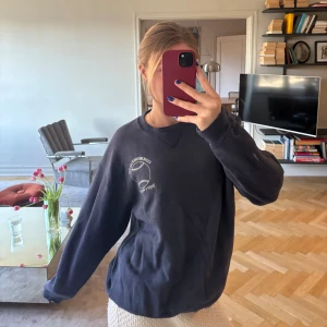 Mörkblå sweatshirt från Commonwear - Säljer en mörkblå sweatshirt från Commonwear med ett tryck av en baseboll och texten 'Umpire Association' på bröstet. Tröjan har långa ärmar och en rund halsringning. Perfekt för en avslappnad stil.