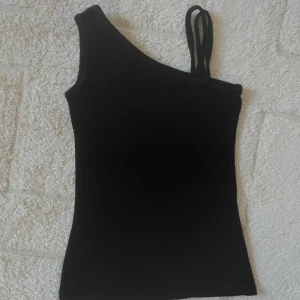 Svart oneshoulder topp -  en svart oneshoulder topp med en axelbandslös design på ena sidan och dubbla smala axelband på andra sidan. 