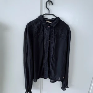 Svart blus med spetsdetaljer - Säljer en svart blus med spetsdetaljer framtill och vid axlarna. Blusen har knappar längs hela framsidan och långa ärmar. Perfekt för dig som gillar en stilren look med lite extra detaljer.