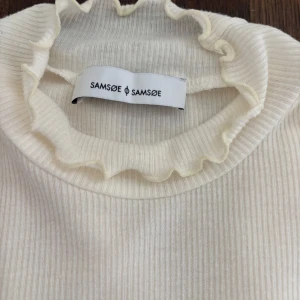 Vit ribbad topp från Samsøe Samsøe - Säljer en vit ribbad topp från Samsøe Samsøe med korta ärmar och hög hals med volangkant. Toppen är figurnära och har en mjuk, stretchig känsla. Perfekt att styla till olika outfits🥰🤗