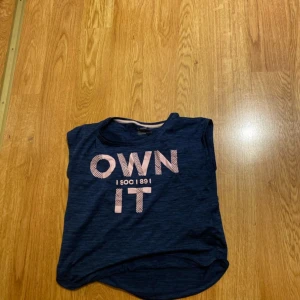 Blå träningstopp från SOC med tryck - Mörkblå träningstopp från SOC med texten 'OWN IT' i rosa framtill. Toppen är ärmlös och tillverkad i ett lätt och luftigt material som passar perfekt för träning eller sportaktiviteter.