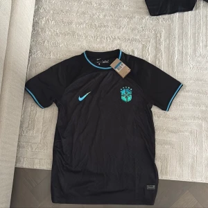 Svart Brasilien fotbollströja från Nike - Säljer en svart Brasilien fotbollströja från Nike med turkosa detaljer på krage, ärmslut och logga. Tröjan har korta ärmar och är tillverkad i ett lätt och ventilerande material. Perfekt för dig som vill sticka ut på planen.