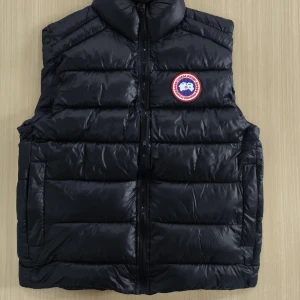 Svart dunväst från Canada Goose - Säljer en svart dunväst från Canada Goose med klassisk logotyp på bröstet. Västen har hög krage, dragkedja framtill och två sidofickor. Perfekt för lager-på-lager under kyliga dagar.