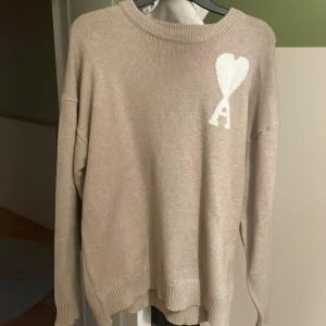 Beige stickad tröja med hjärtdetalj från Ami Paris - Säljer en beige stickad tröja från Ami Paris med en vit hjärtdetalj och bokstaven A på bröstet. Tröjan har rund halsringning och långa ärmar. Perfekt för dig som gillar stilrena och trendiga plagg.