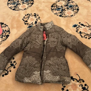 Brun mönstrad dunjacka från Desigual - Säljer en brun dunjacka från Desigual med unikt, detaljerat mönster i beige. Jackan har hög krage och dragkedja framtill. Insidan är dekorerad med blommor och Desigual-logga. Perfekt för dig som vill sticka ut med en snygg och varm jacka.