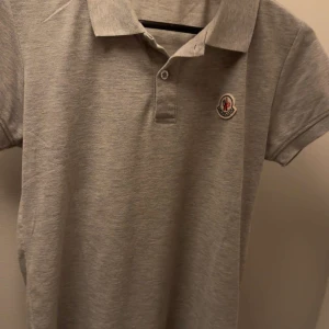 Moncler pike - Säljer en stilren grå pikétröja från Moncler med klassisk krage och knappslå framtill. Tröjan har korta ärmar och en broderad Moncler-logga på bröstet. Perfekt för en avslappnad men snygg look. Använd några gånger inte många alls, priset kan diskuteras.