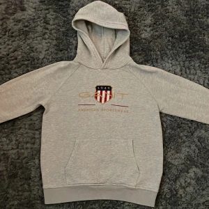 Ljusgrå hoodie från GANT - Ljusgrå hoodie från GANT med broderad logga och sköld på bröstet. Klassisk känguruficka framtill och huva. Mjuk bomullsblandning och ribbade muddar vid ärmslut och nederkant. Perfekt för en avslappnad stil.