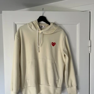 Beige hoodie från Comme des Garçons Play - Säljer en beige hoodie från Comme des Garçons Play med den klassiska röda hjärtloggan på bröstet. Tröjan har huva med dragsko och en stor magficka. Tröjan är i bra skick.