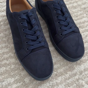 Mörkblå sneakers från Christian Louboutin - Säljer ett par mörkblå sneakers från Christian Louboutin med klassisk röd sula och broderad logga på hälen. Skorna har snörning, rund tå och är tillverkade i mocka med detaljer i skinn på insidan.