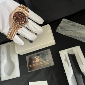 Roséguldfärgad klocka från Nibosi - Snygg klocka i roséguld med metallarmband och mörk urtavla. Klockan har datumvisning och tydliga indexmarkeringar. Perfekt accessoar för dig som vill sticka ut med stilren design och exklusiv känsla.