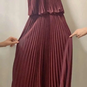 Plisserad vinröd långklänning med gulddetalj - Säljer en elegant vinröd långklänning med plisserat tyg och guldfärgad halterneck-detalj. Klänningen har en lös passform upptill och markerad midja. Perfekt för speciella tillfällen när du vill sticka ut med stil.