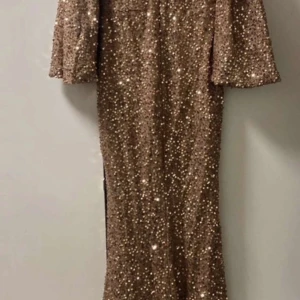 Glittrig guldklänning med trekvartsärm - Säljer en glittrig guldklänning med trekvartsärmar och rak passform. Klänningen är täckt av paljetter och har en elegant lång siluett, perfekt för speciella tillfällen. Supersnygg att styla med klackar och enkla smycken.