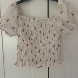 Blommig offshoulder topp med puffärmar från H&M - Supersöt offshoulder topp från H&M i ljusbeige med små blommor och puffiga korta ärmar. Smockad kropp och volangkant nertill. Perfekt för en romantisk look!