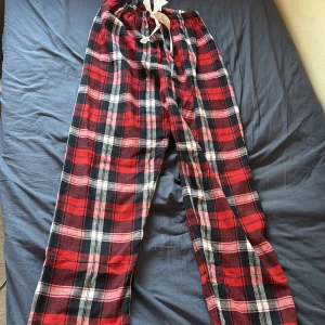 Rödrutiga mjukisbyxor med snörning - Mysiga mjukisbyxor i röd, svart och vit rutig design. Byxorna har resår och snörning i midjan för extra komfort. Perfekta för att chilla hemma eller som pyjamasbyxor. Tillverkade i mjukt syntetmaterial.