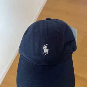 Mörkblå keps från Polo Ralph Lauren - Snygg mörkblå keps från Polo Ralph Lauren med klassisk broderad logga framtill och texten 'POLO' broderad baktill. Justerbart spänne bak för perfekt passform. Perfekt accessoar för en stilren look.