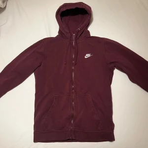 Vinröd hoodie från Nike - Säljer en vinröd hoodie från Nike med dragkedja och huva. Tröjan har två fickor framtill och en vit Nike-logga på bröstet. Perfekt för en avslappnad stil. ( de svarta