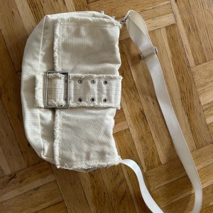 Vit tygväska - Säljer denna tygväska som jag själv har köpt på sellpy. En vit väska i tyg/jeans-material med ett knappstänge och nitar på sidan. Väskan har justerbart band och tecken på användning. Det finns några fläckar (kolla sista bilden) samt att man ser att den är använd💕🌟