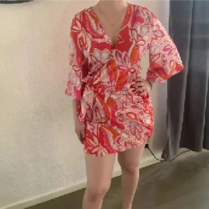 Rosa och röd omlottklänning med blommigt mönster - Säljer en kort zara satin omlottklänning i rosa och rött med blommigt mönster. Klänningen har trekvartsärmar och knyts i midjan för en snygg passform. Perfekt för vår och sommar!