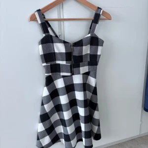 Svartvit rutig klänning med dragkedja - Säljer en svartvit rutig klänning med breda axelband och dragkedja framtill. Klänningen har en utställd kjol och är ärmlös, perfekt för en trendig look. Materialet känns stabilt och mönstret är klassiskt rutigt.