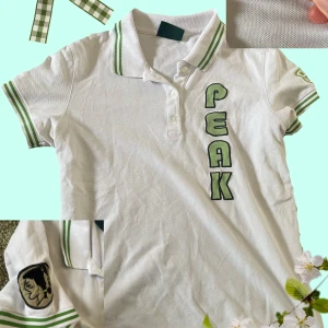 Vit piké t-shirt från Peak - Vit piké t-shirt med gröna ränder på krage och ärmslut. Stora bokstäver 'PEAK' broderade framtill och en liten svart brodyr på ena ärmen. Klassisk krage med knappar och sportig känsla! Andvänt en gång, inga fläckar. 