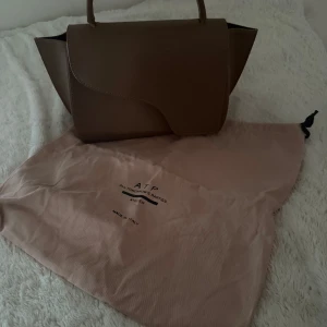Brun handväska från ATP Atelier - Säljer en stilren brun handväska från ATP Atelier med elegant form och minimalistisk design. Väskan har ett topphandtag och är tillverkad i slätt skinn. Perfekt för dig som gillar klassiska accessoarer med modern touch. Det ingår även ett långt band till väskan. Äkta!!💕