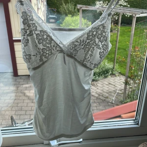 Vit spetslinnetopp med blommönster - Säljer en vit linnetopp och vacker spets upptill. Toppen har blommönstrad spets över bysten och en liten rosett framtill. Perfekt för lager-på-lager eller sommarens varma dagar.