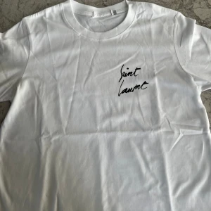 Saint Laurent t-shirt - Säljer en vit t-shirt från Saint Laurent med svart tryck på bröstet och stor logga på ryggen. Klassisk rund hals och korta ärmar. Perfekt för dig som gillar stilrena och trendiga plagg med designer-touch.