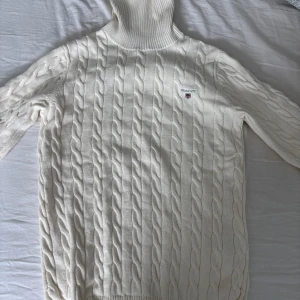 Vit kabelstickad polotröja från GANT - Säljer en klassisk vit kabelstickad polotröja från GANT med broderad logga på bröstet. Tröjan har lång ärm, hög polokrage och ribbade muddar. Perfekt för kyliga dagar och enkel att matcha med olika outfits.