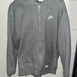 Grå hoodie med dragkedja från Nike - Klassisk grå hoodie från Nike med dragkedja framtill och vit logga på bröstet. Tröjan har huva med dragsko och två fickor framtill. Perfekt för en avslappnad stil.