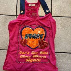 Rosa linnetopp med paljetter och spets från Kookai - Unik rosa linnetopp med stora hjärtan, paljetter och texten 'Let's Do The Time Warp Again' framtill. Axelband i denim och baksida i genomskinlig spets med blommönster. Perfekt för dig som vill sticka ut! Storlek XS/S