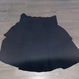 Svart volangkjol med smock i midjan - Supersöt svart kjol med volanger och smockad midja. Kjolen är kort och har en luftig känsla, perfekt för varma dagar. Passar till många olika stilar och är enkel att matcha med olika toppar.