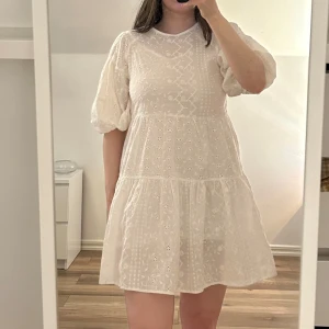 Vit smockklänning med broderi - Superfin vit kortklänning med puffärmar och broderade blommor. Klänningen har en lös passform, rund halsringning och volang nertill. Har en tunn underklänning. Oanvänd. 