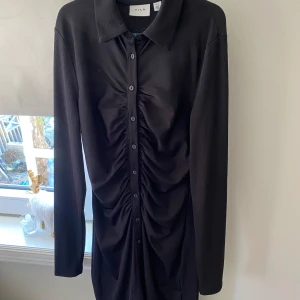 Svart skjortklänning med rynk från Vila - Svart skjortklänning från Vila med långa ärmar och knappar framtill. Klänningen har snygga rynkdetaljer längs knappraden och klassisk krage. Perfekt för dig som gillar stilrena och trendiga plagg. Kom med bud!