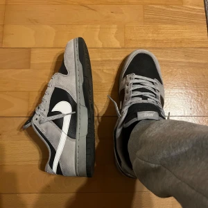 Nike Dunk Low i svart och grå mocka - Säljer ett par Nike Dunk Low med svart bas och gråa paneler i mocka. Skorna har vit swoosh, grå snörning och svart platt sula. Perfekta för dig som gillar klassisk streetwear-stil.