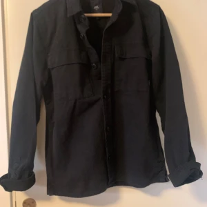 Svart overshirt lindbergh - Säljer en svart overshirt med klassisk krage och knäppning framtill. Skjortan har långa ärmar och två bröstfickor med lock. Perfekt att bära som lager över en t-shirt eller tröja. Tröjan är perfekt nu för sommaren o vinter. Ny pris 1200 mitt pris 349