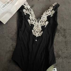 Svart bodysuit med spetsdetaljer från Intimissimi - Säljer en svart bodysuit med djup v-ringning och vackra vita spetsdetaljer framtill. Perfekt för att känna sig extra fin! Materialet är mjukt och följsamt.