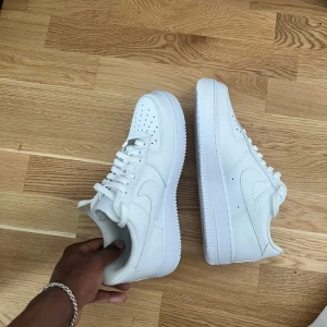 Nike Air Force 1 vita sneakers - Klassiska helvita Nike Air Force 1 sneakers med rund tå och platt sula. Skorna har snörning och är tillverkade i syntetmaterial med diskreta Nike-loggor på sidorna. Perfekta för en clean och stilren look. HELT NYA!