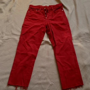 Röda vintage jeansbyxor med råa benslut - Röda jeansbyxor från H&M i vintage fit med raka ben och råa, fransiga benslut. Byxorna har knappar i gylfen och klassiska fem fickor. Liten i storleken. Slitna detaljer på fickor 