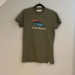 Olivgrön t-shirt med tryck från Topo Designs - Olivgrön t-shirt med ett tryck av en blå bil med röd kanot på taket och texten 'No Fixed Destination' på bröstet. 